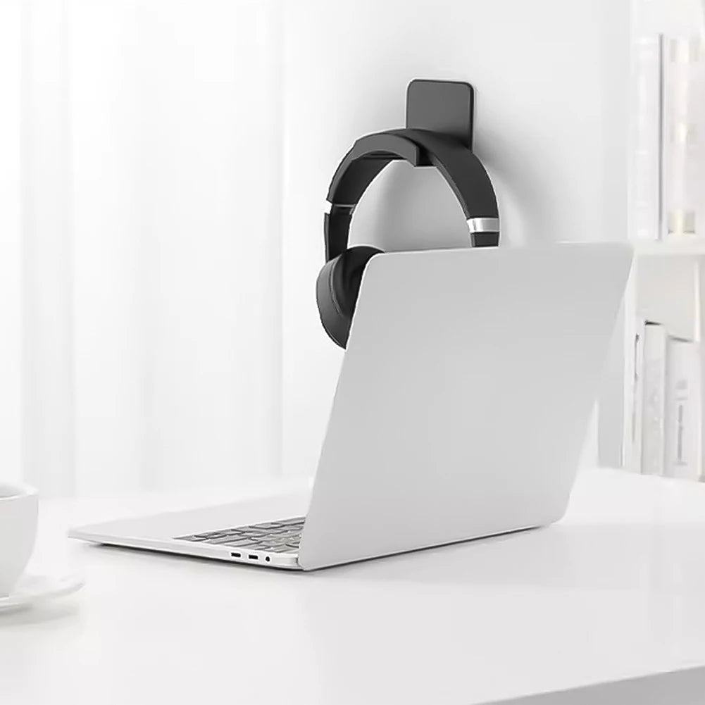 Black headphone hanger mounted on a white wall next to a laptop on a desk, showcasing a sleek and organized workspace.
