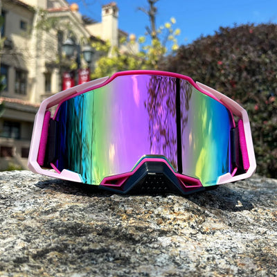 Colorful motorcycle and MTB goggles resting on a rock, showcasing a rainbow mirror lens against a blurred background.