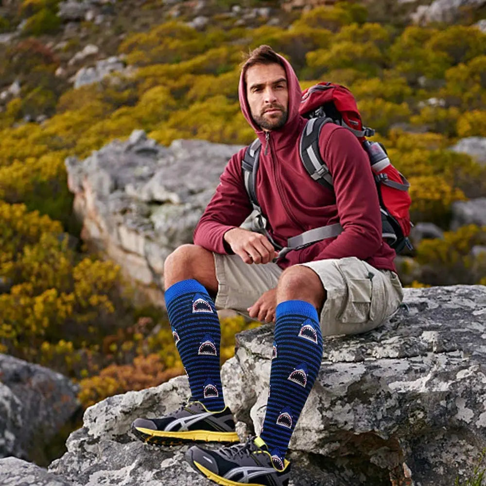 Man wearing blue patterned knee-high socks and hiking gear sitting on a rock in nature.