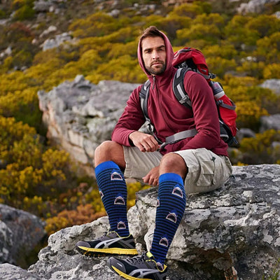 Man wearing blue patterned knee-high socks and hiking gear sitting on a rock in nature.