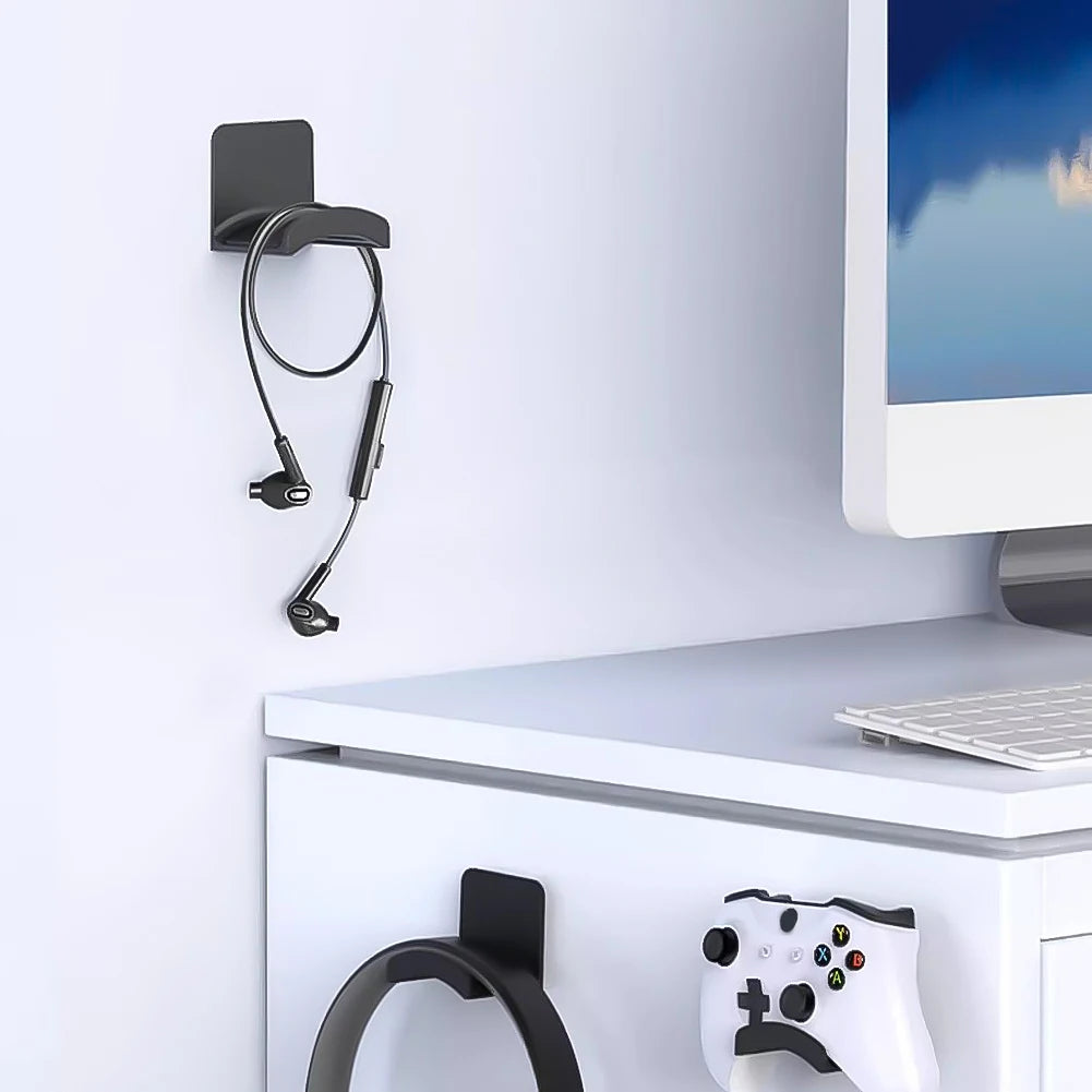 Universal adhesive headphone hanger mounted on wall, holding headset beside a gaming console and desk with computer.